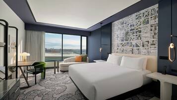 A modern hotel room with a large bed, a desk with a chair, a sofa, and a cityscape view through the window.