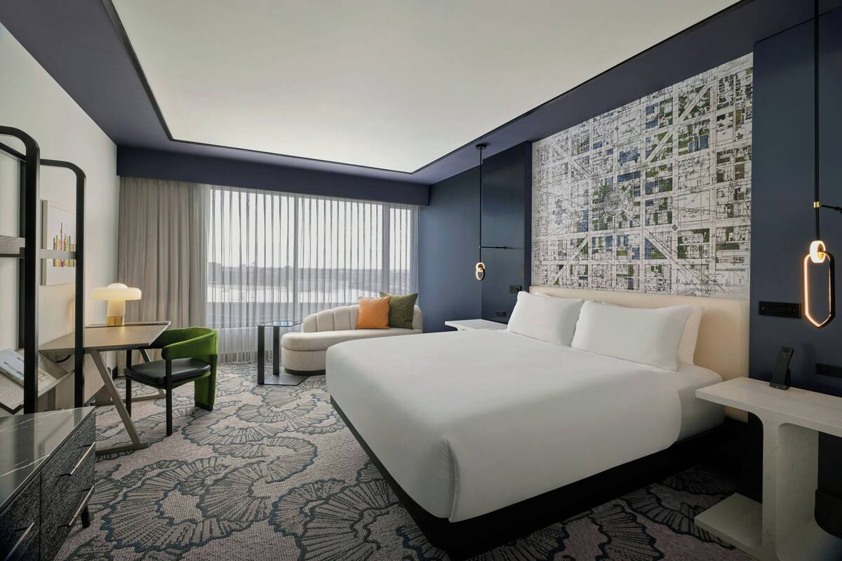 A modern hotel room with a large bed, a desk with a chair, a sofa, and a large window with blinds.