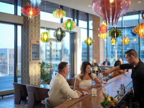 Eine Bar mit Blick auf die Stadt durch große Fenster, ein Barkeeper serviert Getränke, und bunte Hängelampen.