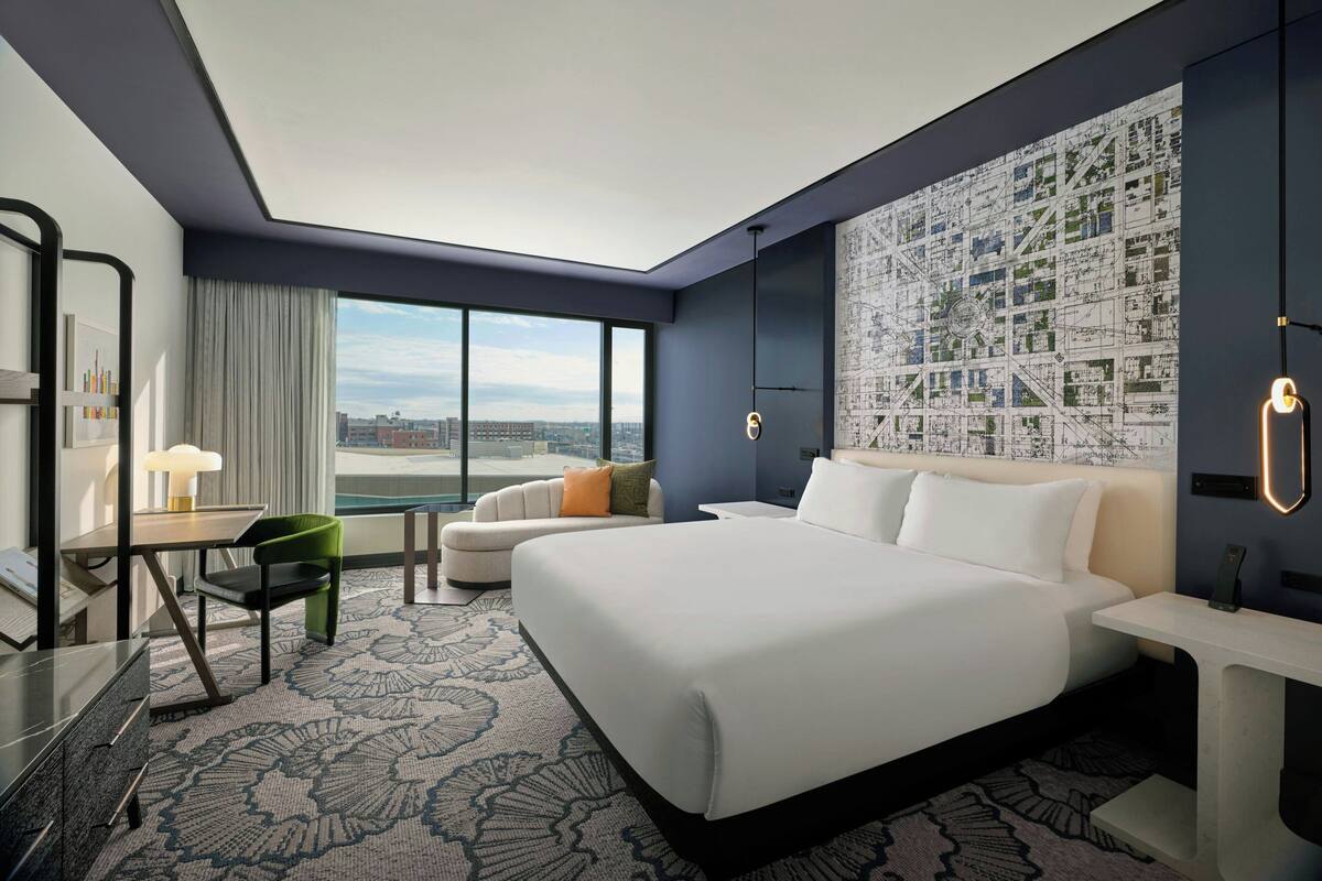 A modern hotel room with a large bed, a desk with a lamp, a chair, and a view of the cityscape.
