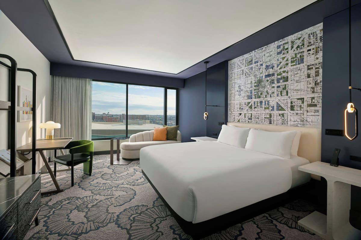 Habitación de hotel moderna con una cama grande, un escritorio con lámpara, una silla y vistas a la ciudad.