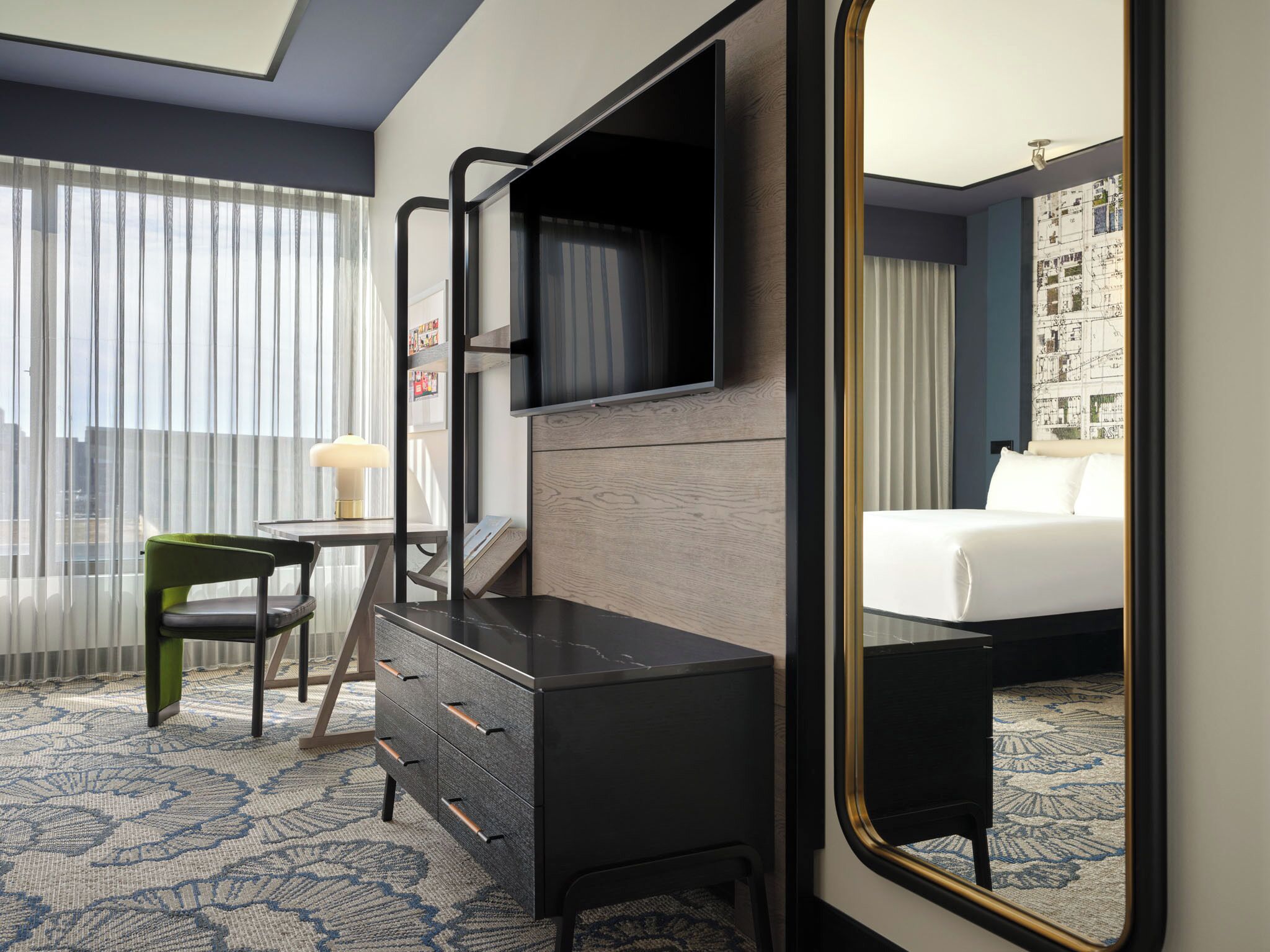 Een hotelkamer met een bed, een bureau met lades, een stoel, een aan de muur bevestigde televisie en een grote spiegel.