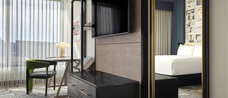 Habitación de hotel con cama, escritorio con cajones, silla, televisor montado en la pared y un espejo grande.