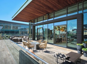 A modern outdoor seating area with wicker chairs and tables, large glass windows, and a view of the cityscape. - The Alexander, Autograph Collection (Indianapolis)