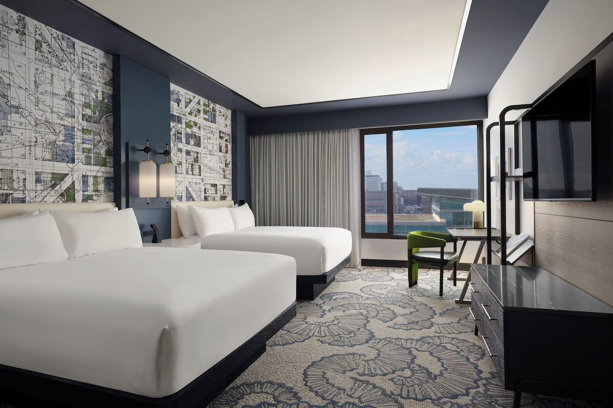 A hotel room with two beds, a desk, a chair, a TV, and a large window with a city view.