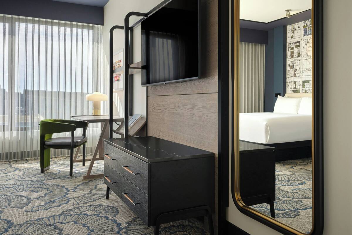 A hotel room with a bed, a desk with drawers, a chair, a television, and a large mirror.