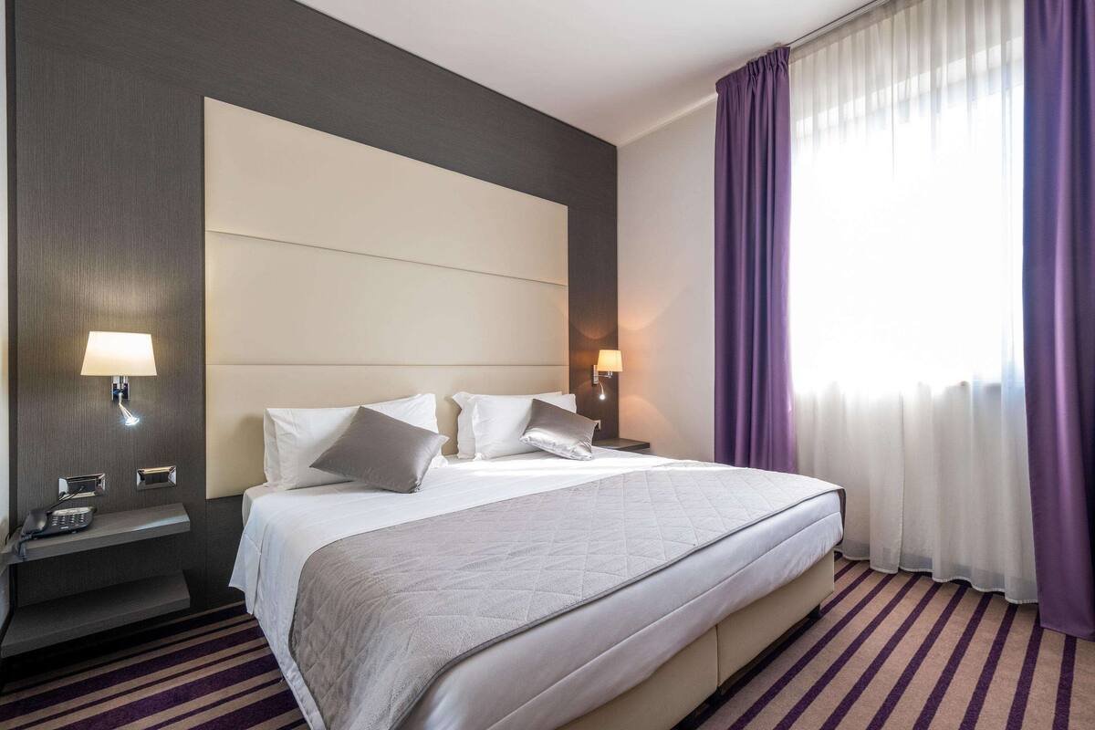 A hotel room with a large bed, two bedside tables with lamps, a dark wall panel, and a window with purple curtains.