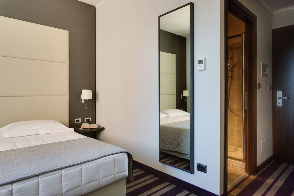 A hotel room with a bed, a nightstand, a mirror, a door to a shower area, and a wall-mounted thermostat.