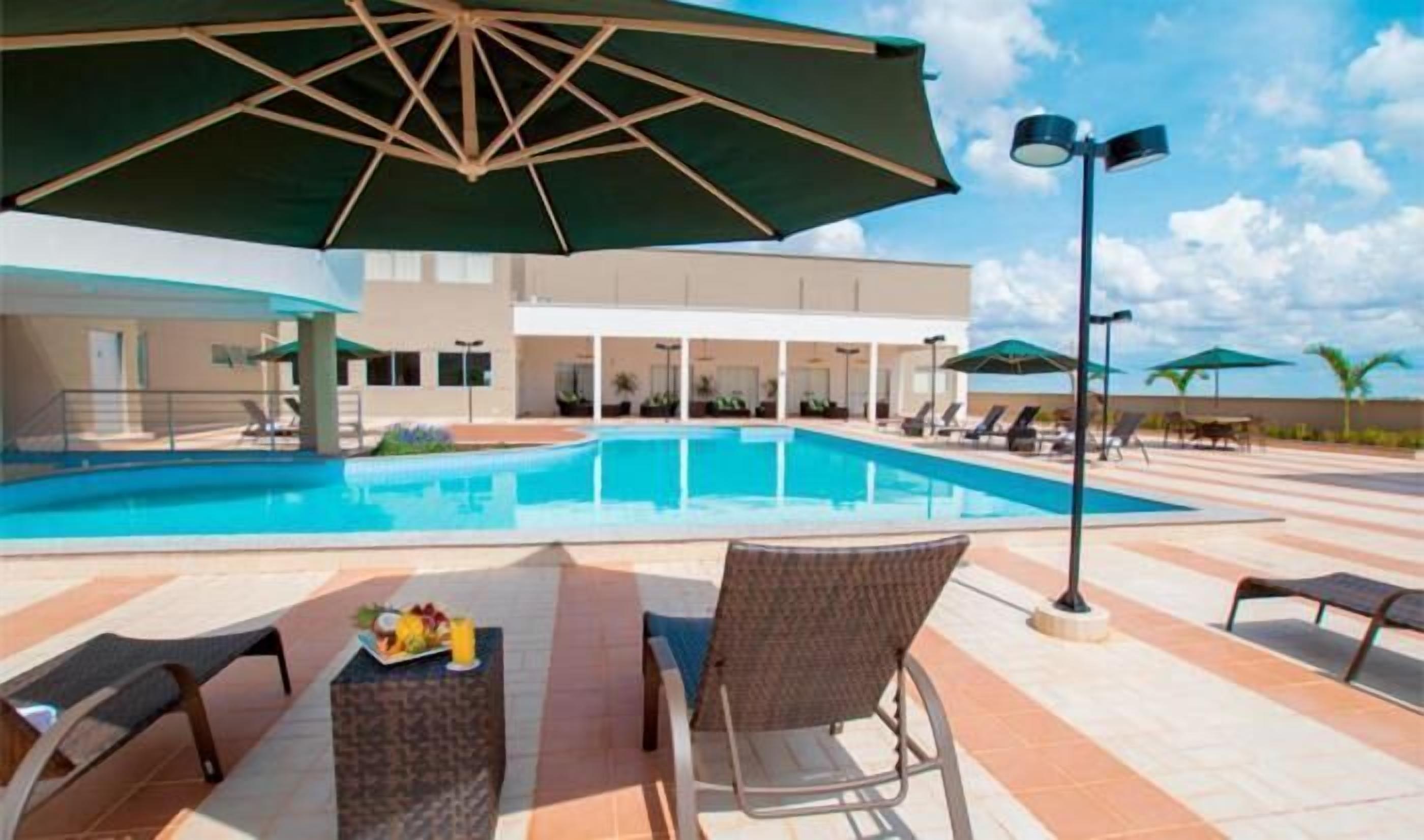 A swimming pool with lounge chairs and umbrellas, surrounded by a building with multiple windows and a clear sky.