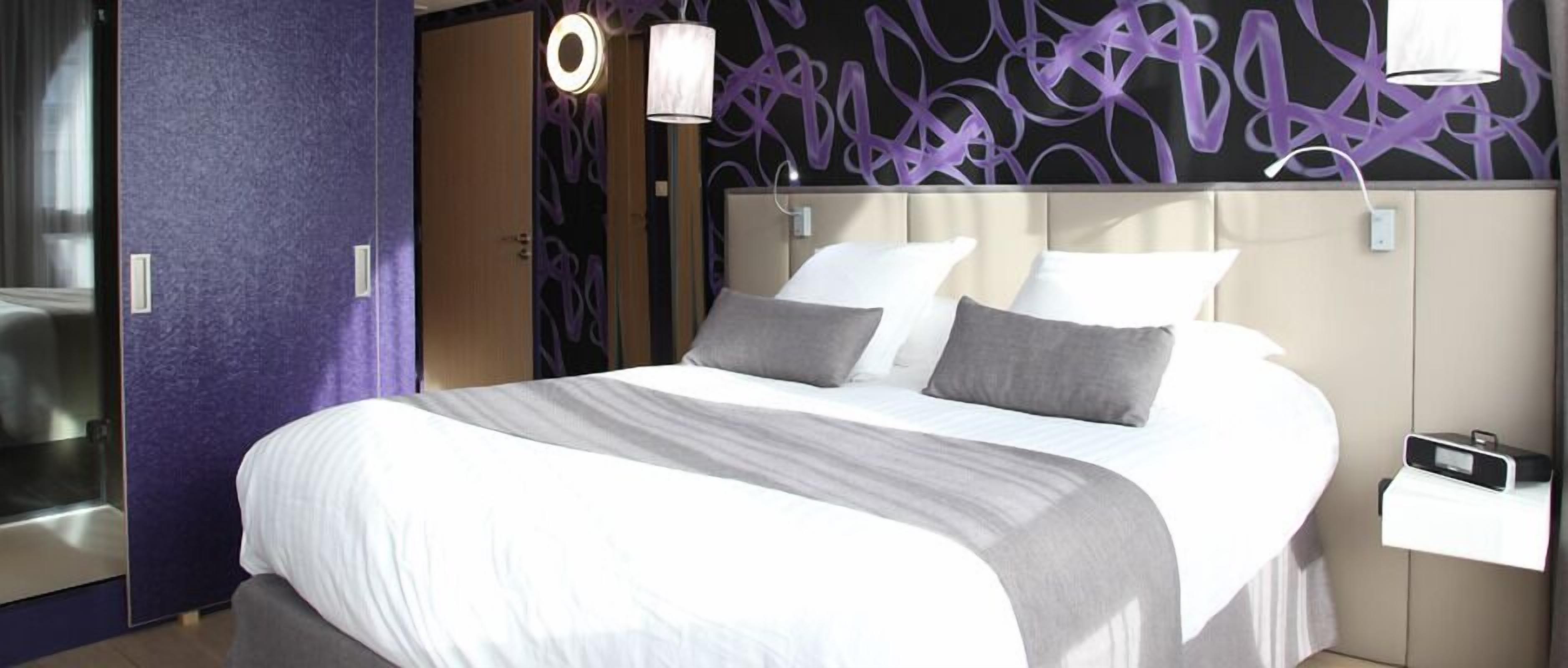 Une chambre d’hôtel moderne dotée d’un grand lit, d’une tête de lit avec éclairage intégré et d’un motif mural abstrait aux tons violets.