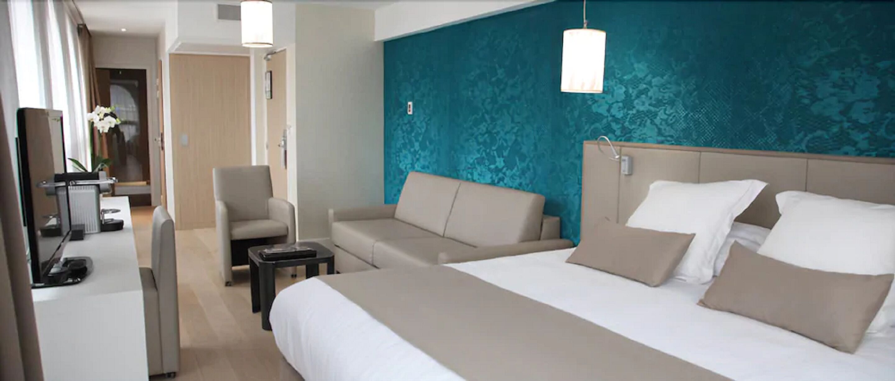 Habitación de hotel con una cama grande, dos sillones, un escritorio con una computadora y una pared de acento color verde azulado.