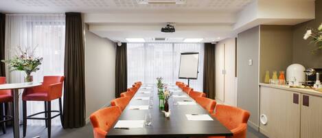 A conference room with a long table, orange chairs, a projector, a whiteboard, and a small kitchenette.