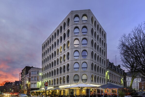 A modern building with a unique geometric facade, featuring multiple circular windows and a ground-level cafe area with outdoor seating. - Best Western Premier Why Hotel (Lille)