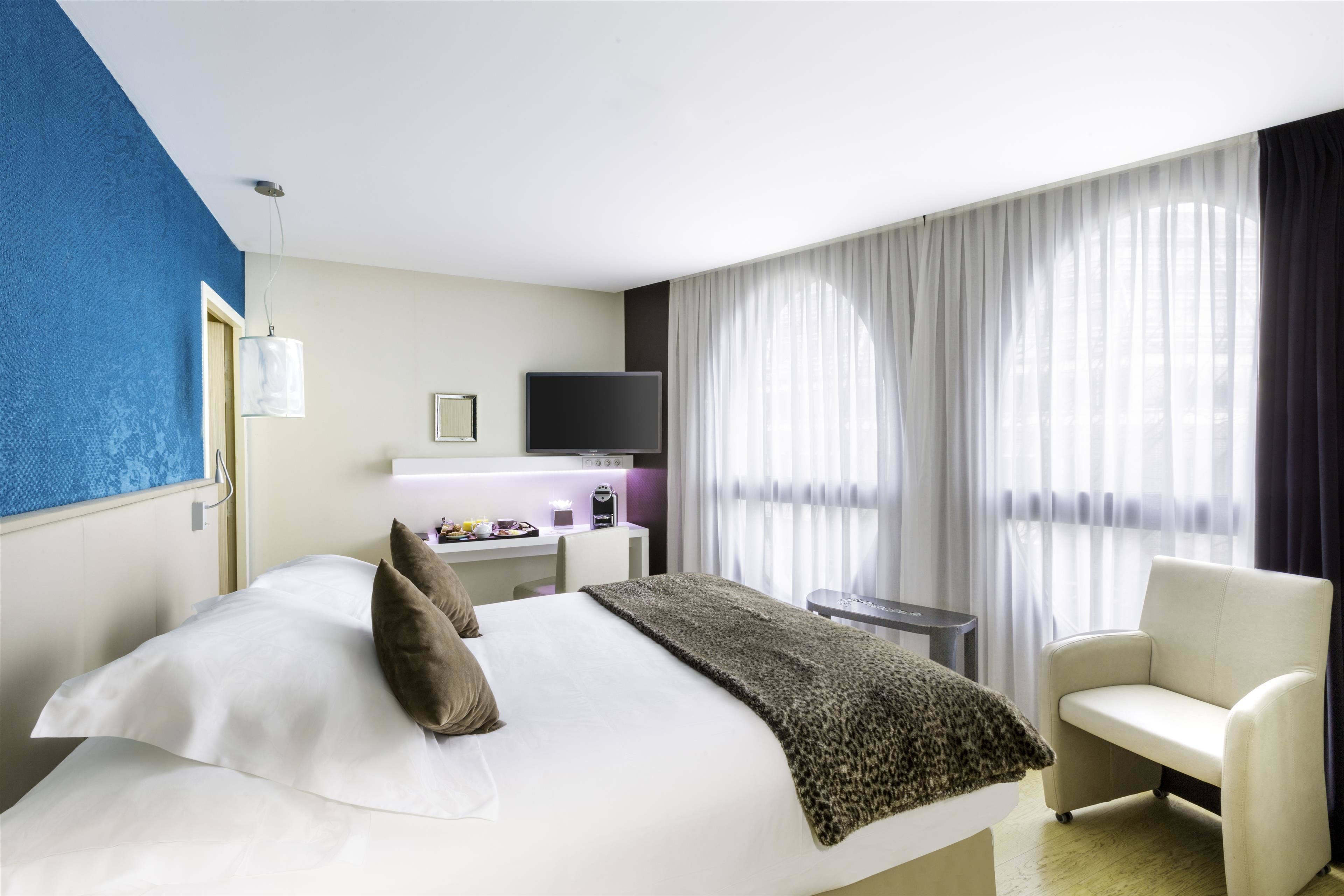 A hotel room with a bed, a chair, a desk with a computer, a TV, and a window with curtains.