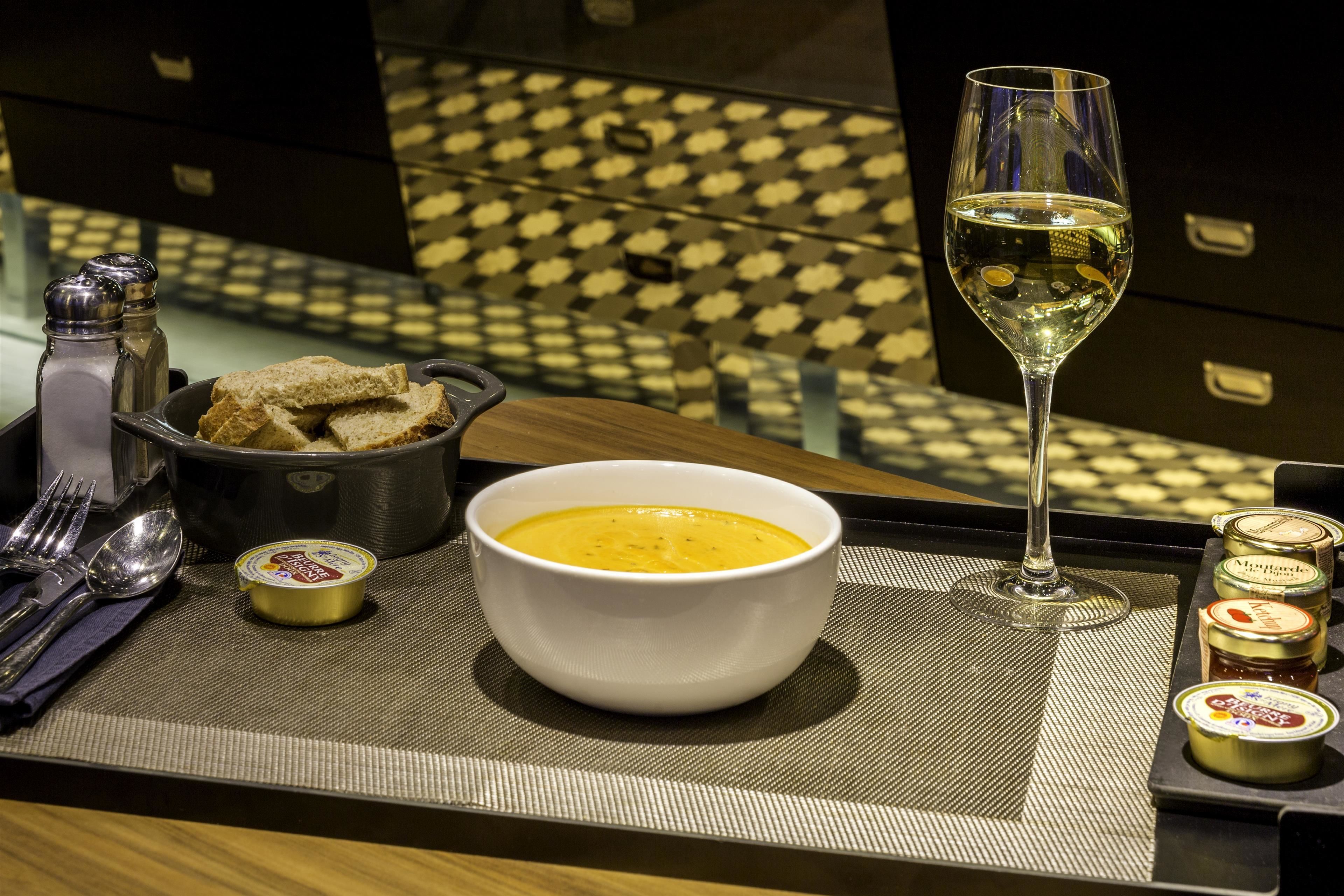 A dining table with a bowl of soup, a glass of white wine, bread, condiments, and cutlery.