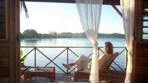 A room with a bed, a wooden table, and a chair facing a lake. - L'Heure Bleue (Nosy Be)