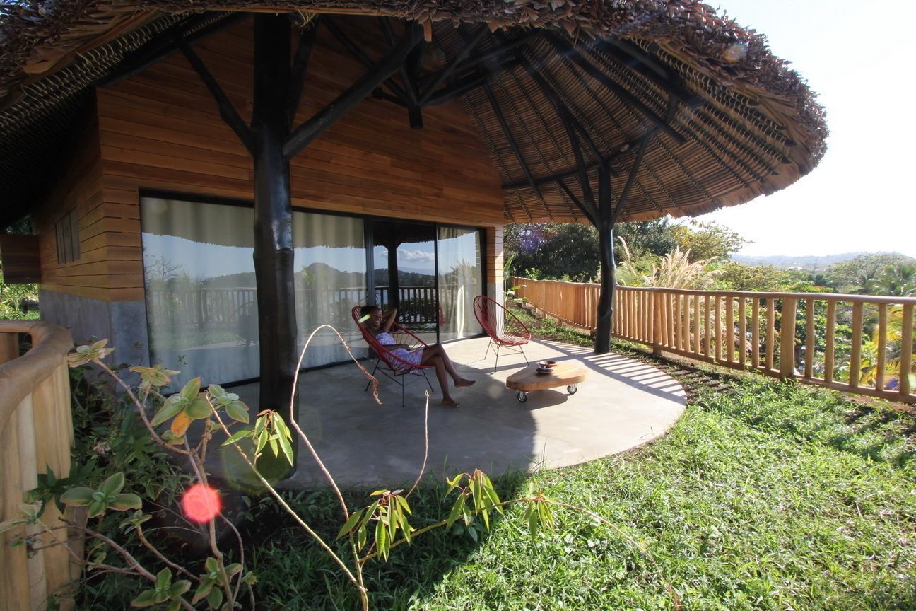 Eine Holzhütte mit überdachter Veranda, einer Glastür, die auf eine Terrasse führt, und Blick auf die umliegende Landschaft.