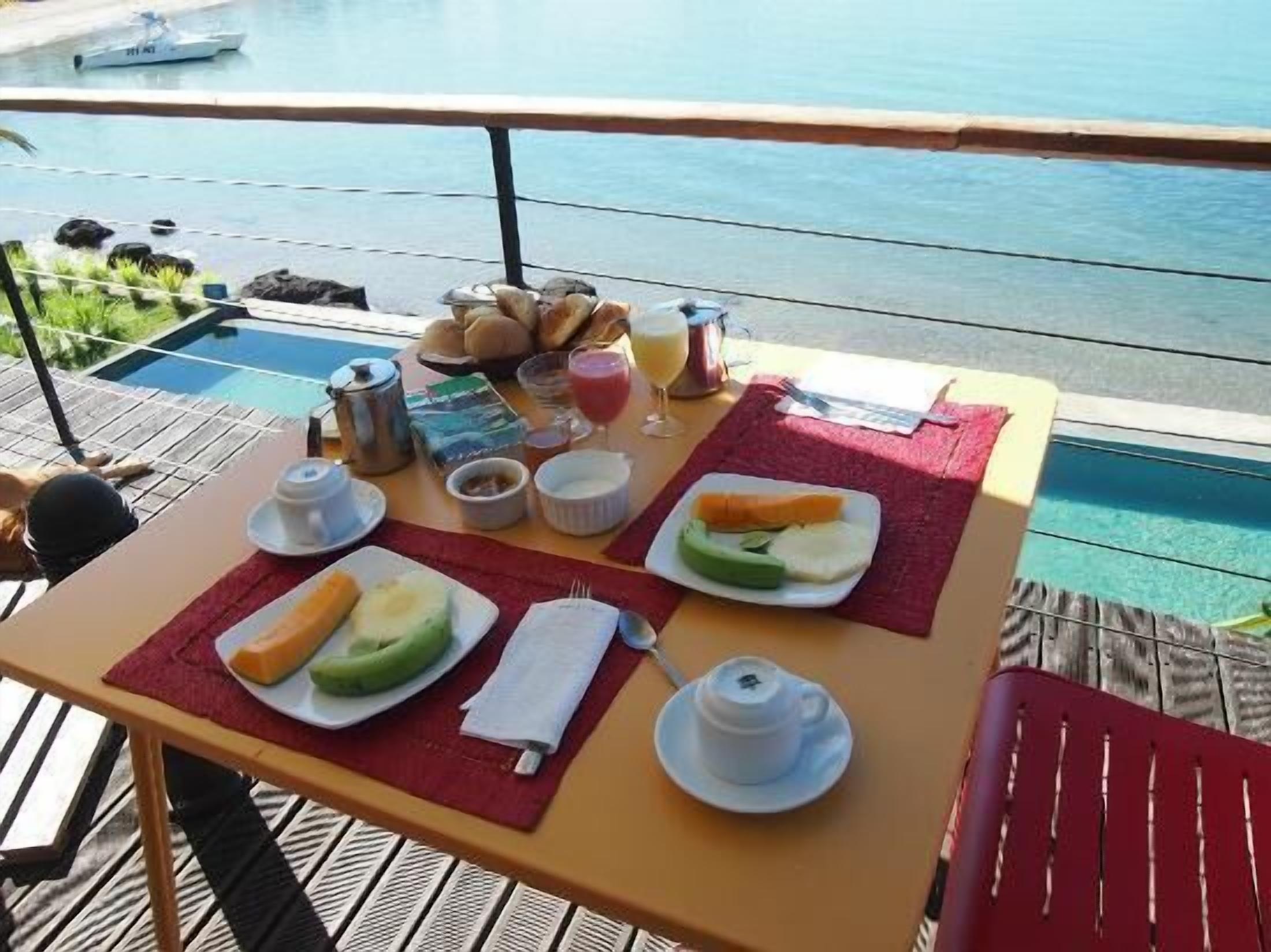 A table with breakfast items including bread, fruit, and beverages, overlooking a pool and ocean view.