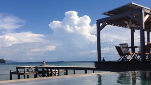 A pier extending into a calm body of water with a clear sky. There are lounge chairs and a covered seating area. - L'Heure Bleue (Nosy Be)