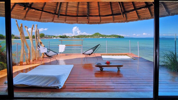 A wooden deck with two lounge chairs and a small table, overlooking a clear blue sea and a distant island. - L'Heure Bleue (Nosy Be)