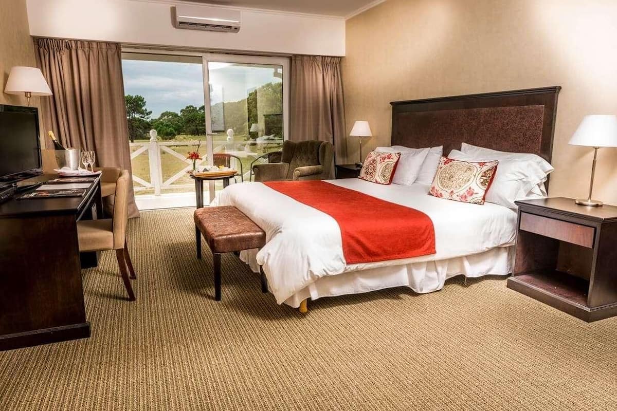 A hotel room with a large bed, a desk, a chair, a TV, and a balcony with curtains.