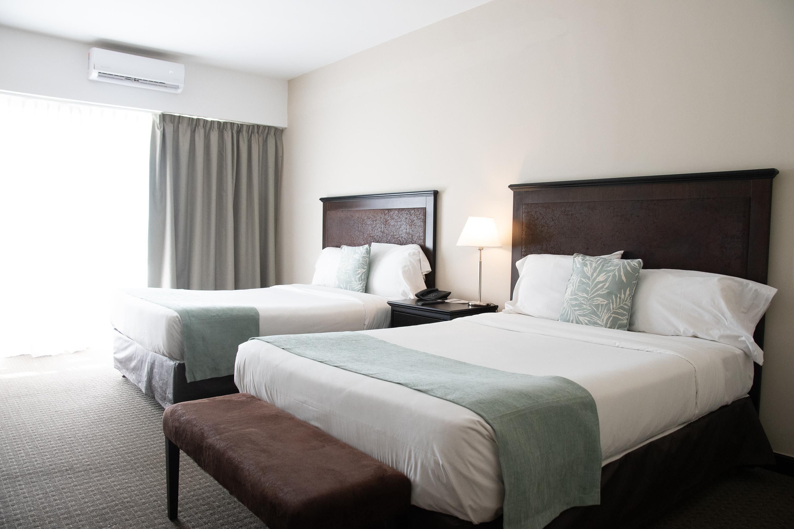 Habitación de hotel con dos camas, un banco, una mesita de noche con lámpara y un equipo de aire acondicionado.
