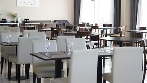 Zona pranzo con diversi tavoli e sedie, un lampadario e ampie finestre con tende.