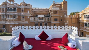 A bed with red bedding and white pillows in front of a large, ornate building with multiple domes and balconies. - Deogarh Mahal (Devgarh)