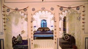 A room with a bed, a small table, and a chair. There are curtains and a window with a view of the outside. - Deogarh Mahal (Devgarh)