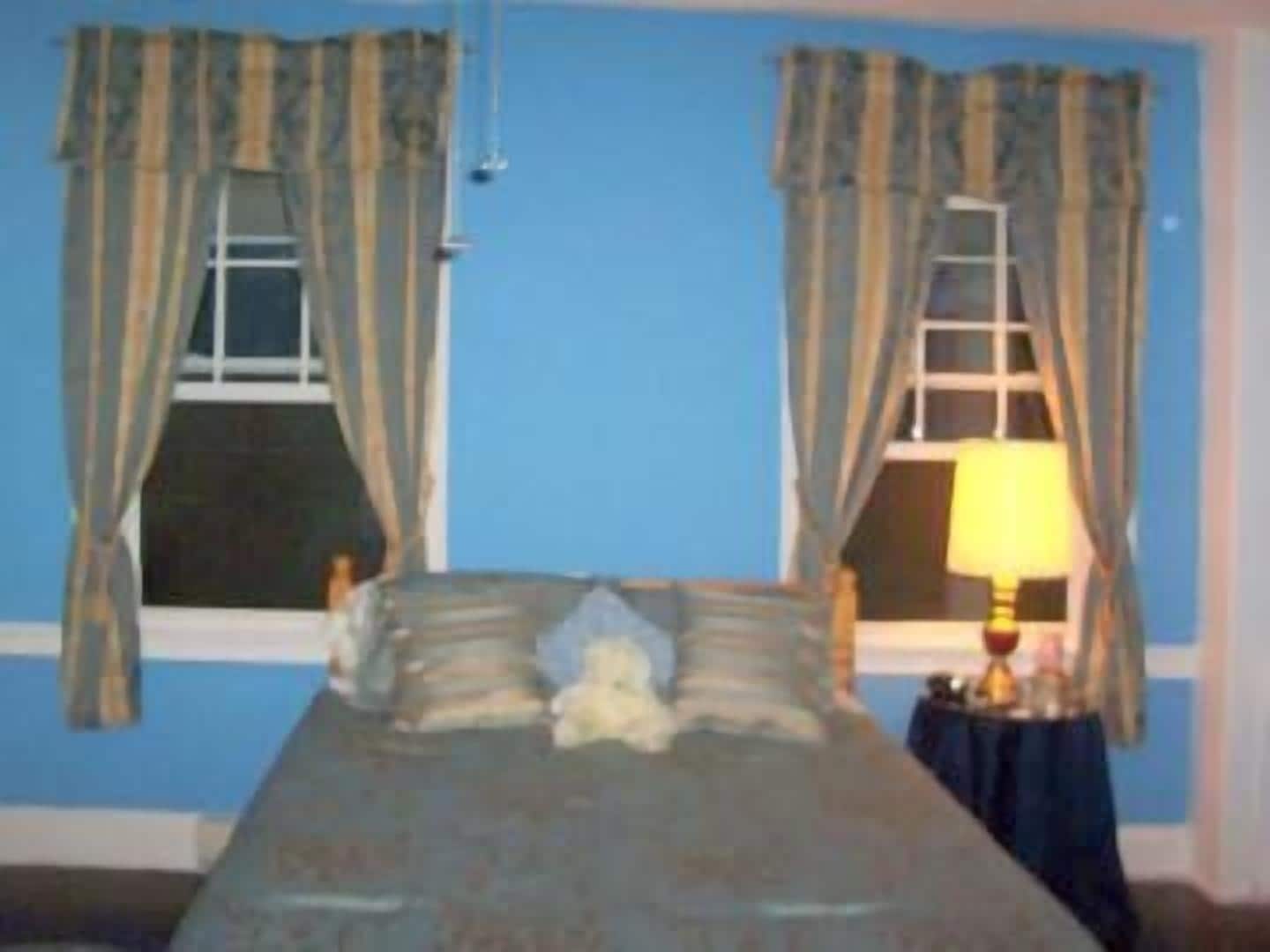 Une chambre à coucher avec un lit, deux fenêtres avec des rideaux, une table de chevet avec une lampe et une échelle appuyée contre le mur.