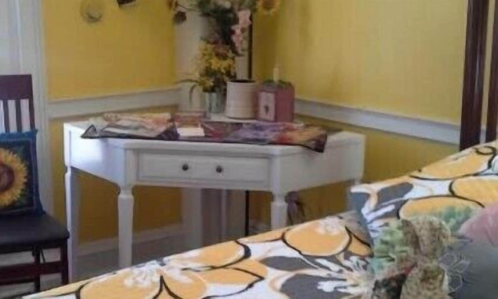 Um quarto com parede amarela, uma mesa branca com gavetas, uma cadeira com almofada de girassol e uma cama com colcha estampada.