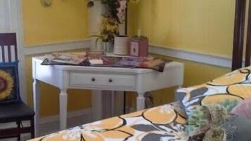Um quarto com parede amarela, uma mesa branca com gavetas, uma cadeira com almofada de girassol e uma cama com colcha estampada.