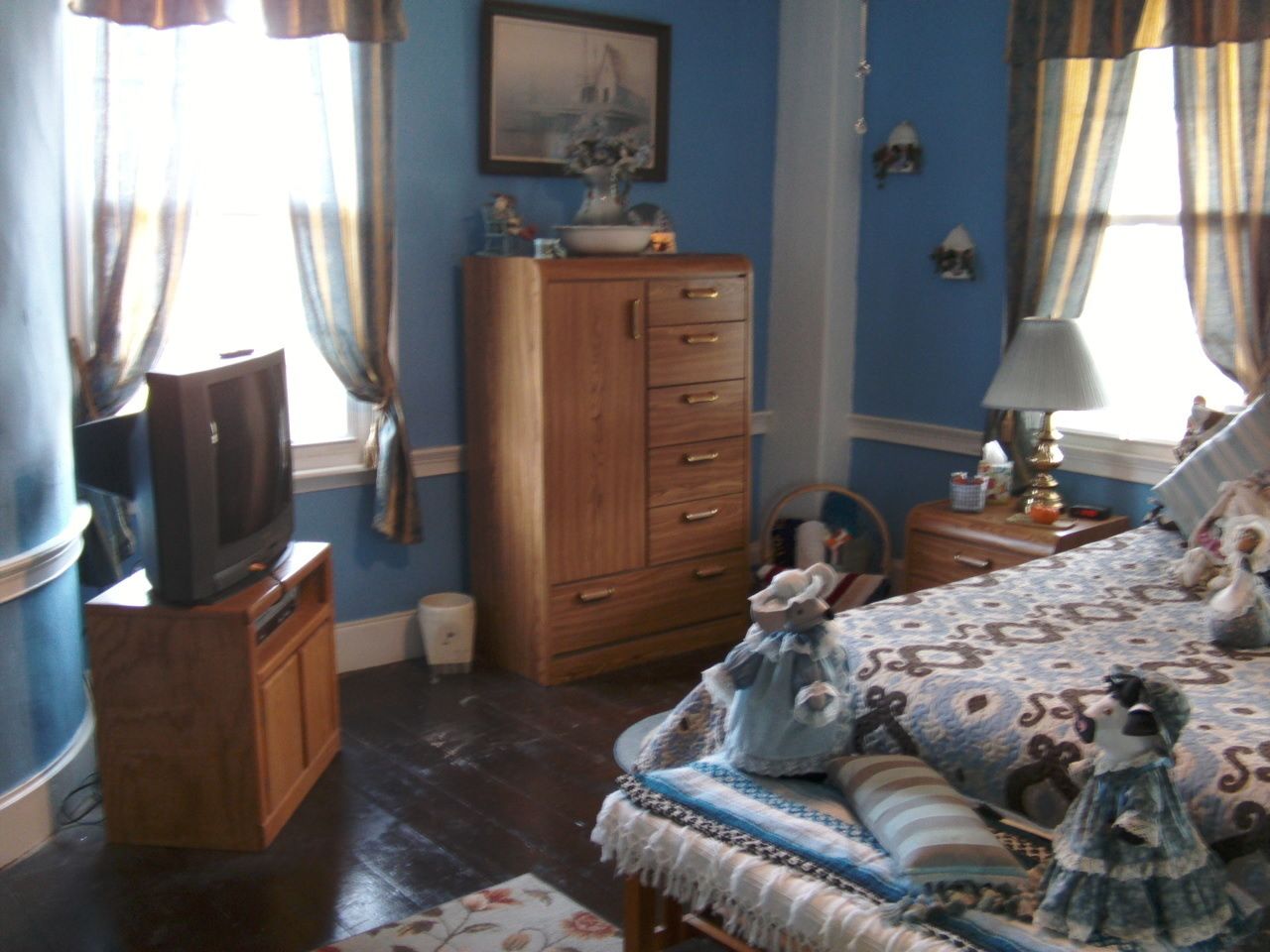 A bedroom with a wooden dresser, a television on a stand, a bed with a floral bedspread, and a nightstand with a lamp.