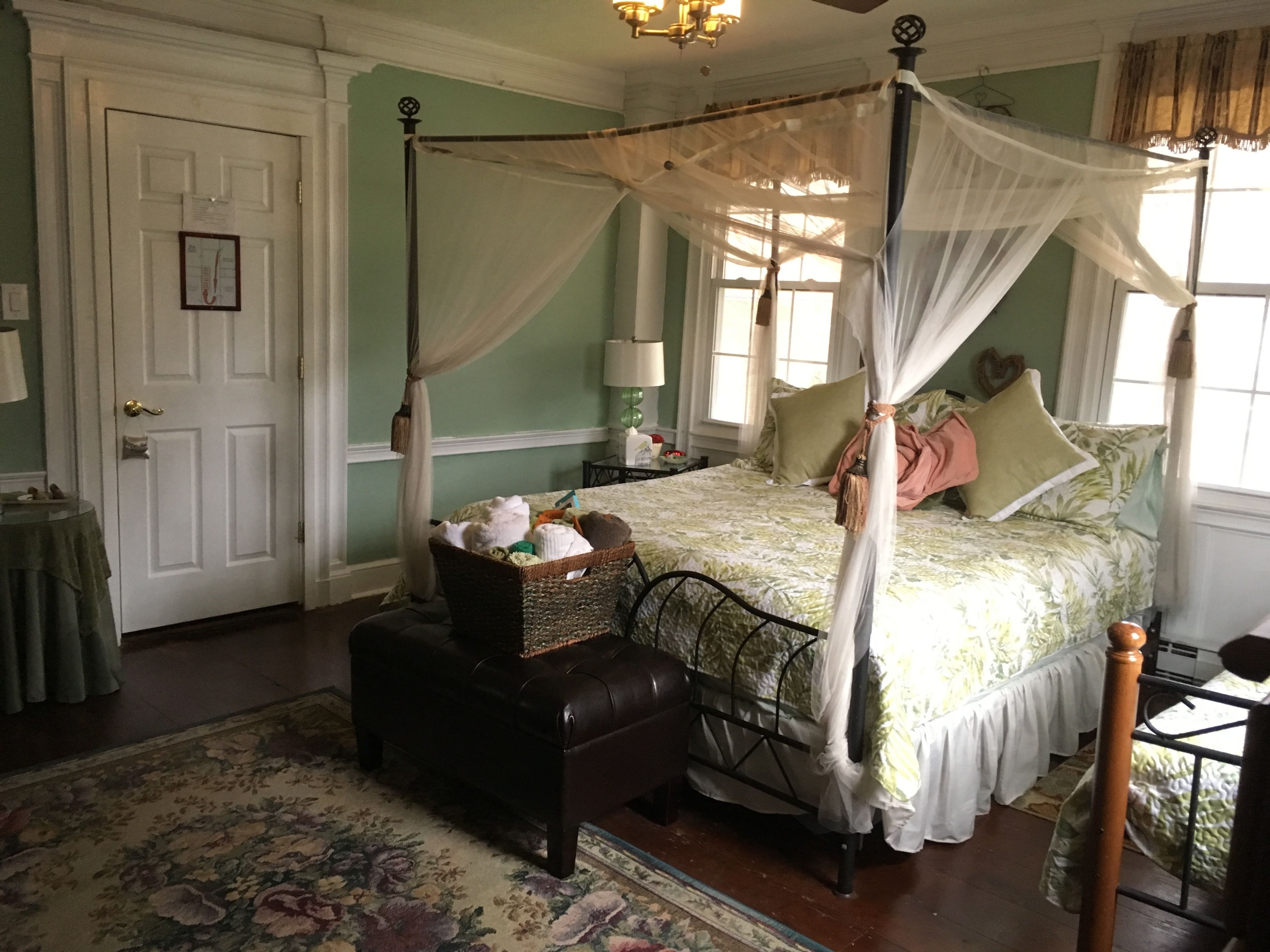 Un lit à baldaquin à quatre poteaux, surmonté d’une couvre-lit à motifs floraux, une table de chevet avec une lampe et une commode en osier.