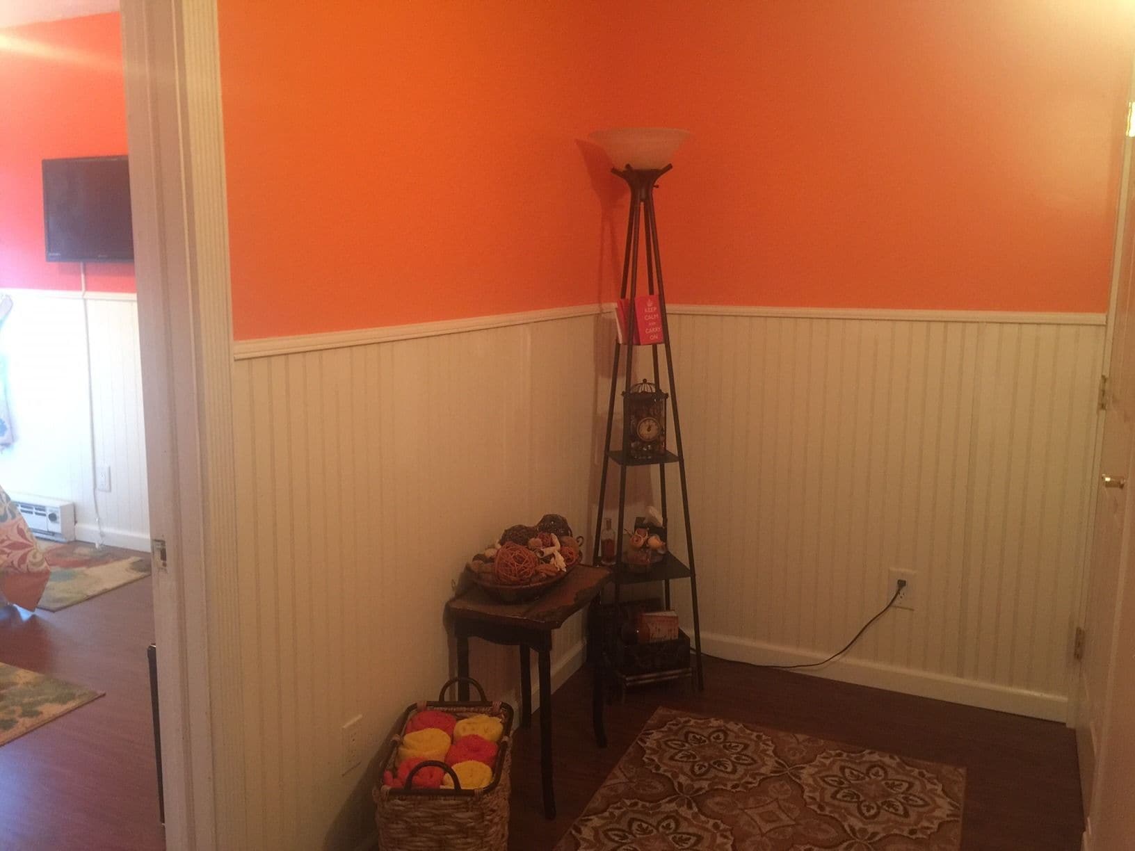 Une pièce aux murs orange, avec une porte blanche, un tapis à motifs, une petite table sur laquelle se trouve un panier, et un grand lampadaire.