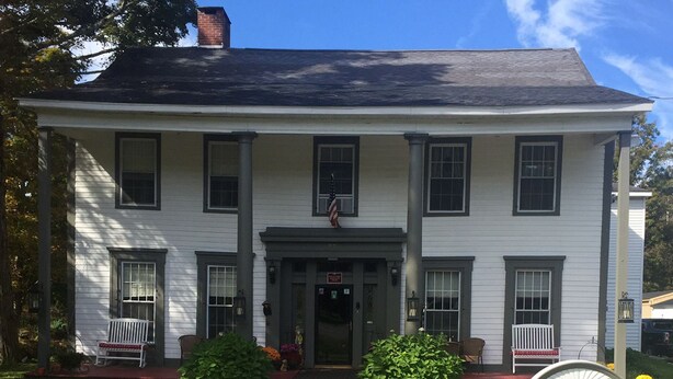 A two-story white building with a central entrance, two front porches with white benches, and a small American flag above the entrance.