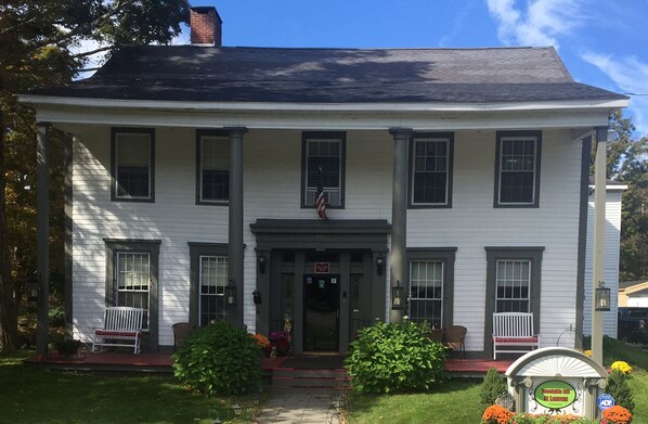A two-story white building with a central entrance, two front porches with white benches, and a small American flag above the entrance. - Brookside Inn At Laurens (Laurens)