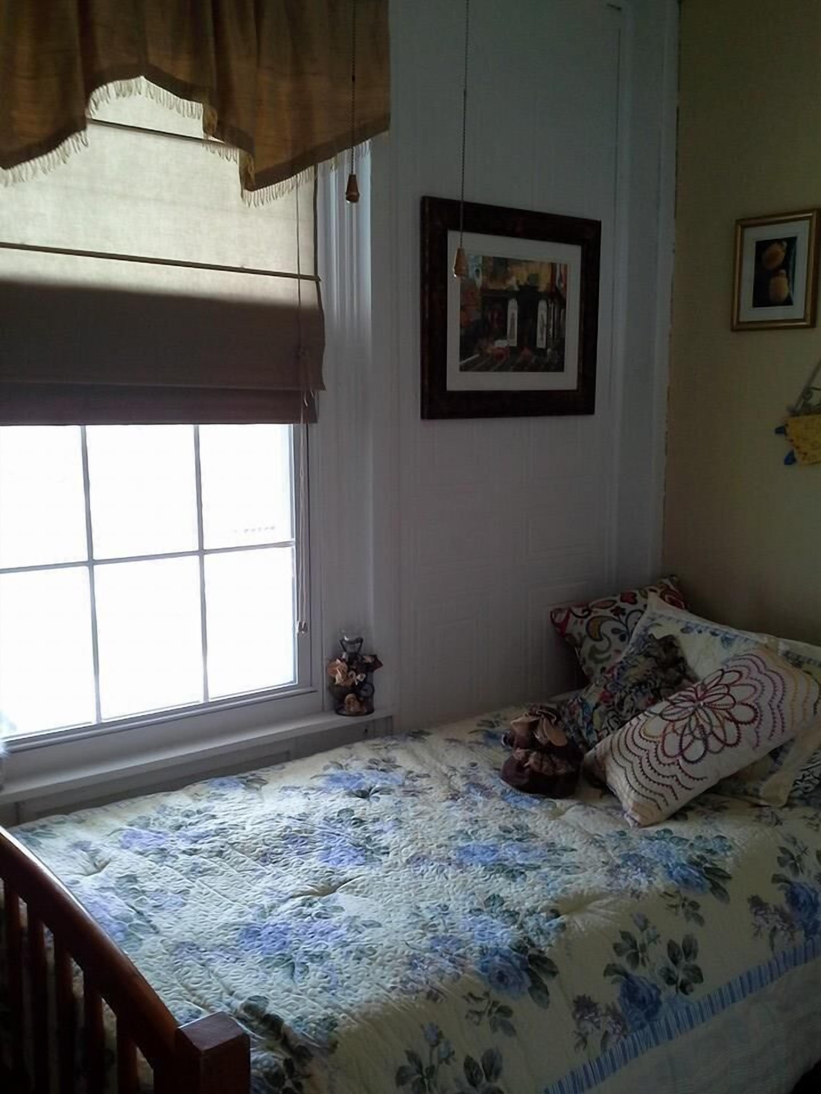 Une chambre à coucher avec un lit, une fenêtre, un tableau encadré et une petite table sur laquelle est posé un objet décoratif.