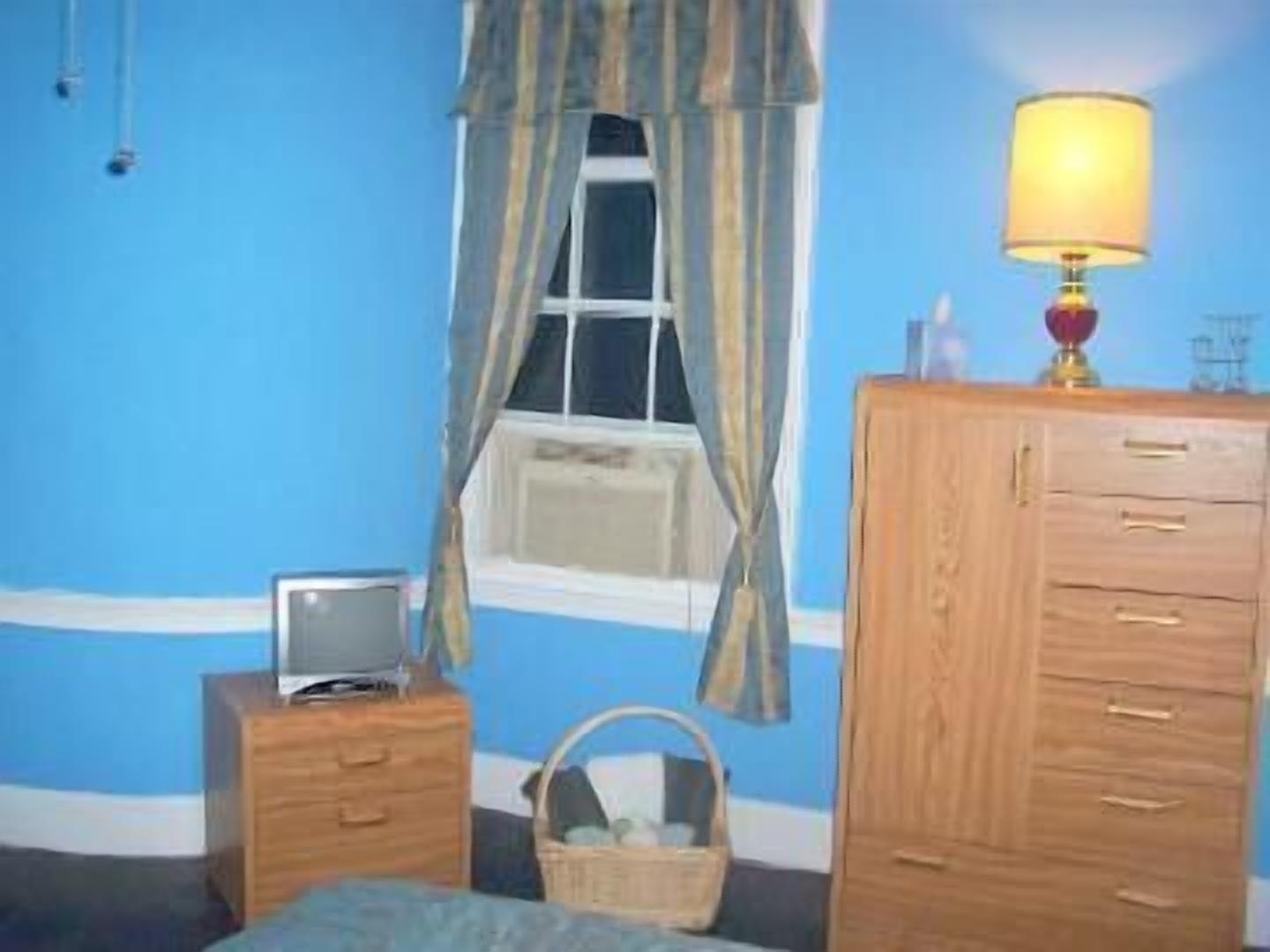 A bedroom with a blue wall, a wooden dresser, a bedside table with a lamp, and a window with curtains.