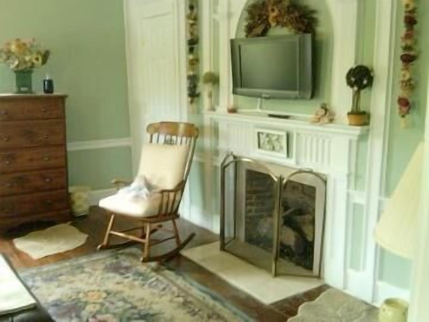 A room with a wooden dresser, a rocking chair, a fireplace, and a television mounted above it.