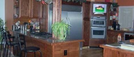 A kitchen with wooden cabinets, a central island, bar stools, a refrigerator, an oven, and a TV mounted on the wall.