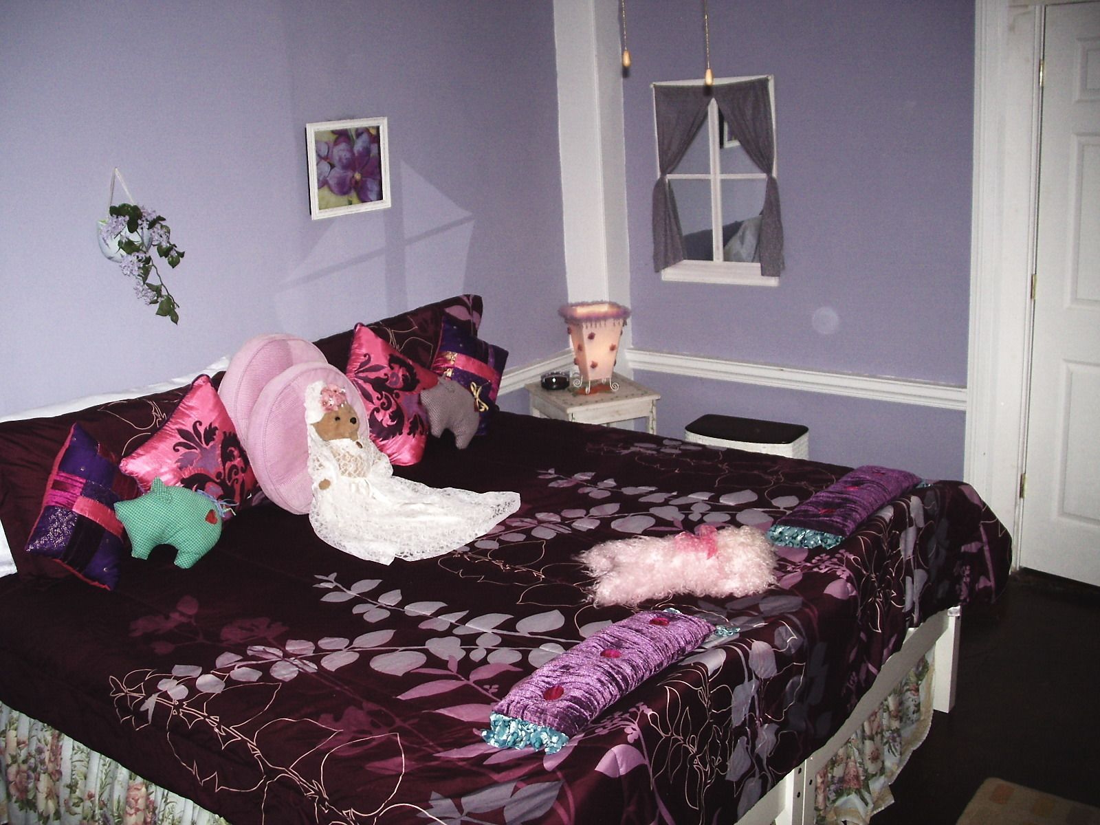 A bedroom with a bed, pillows, a hanging plant, a nightstand, and a window.