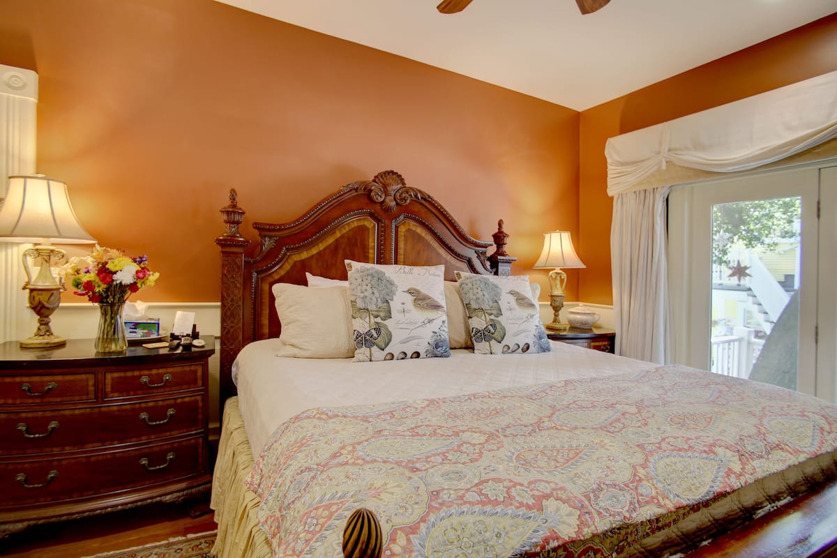 A bedroom with a large bed, ornate headboard, two bedside tables with lamps, a dresser, and a window with curtains.