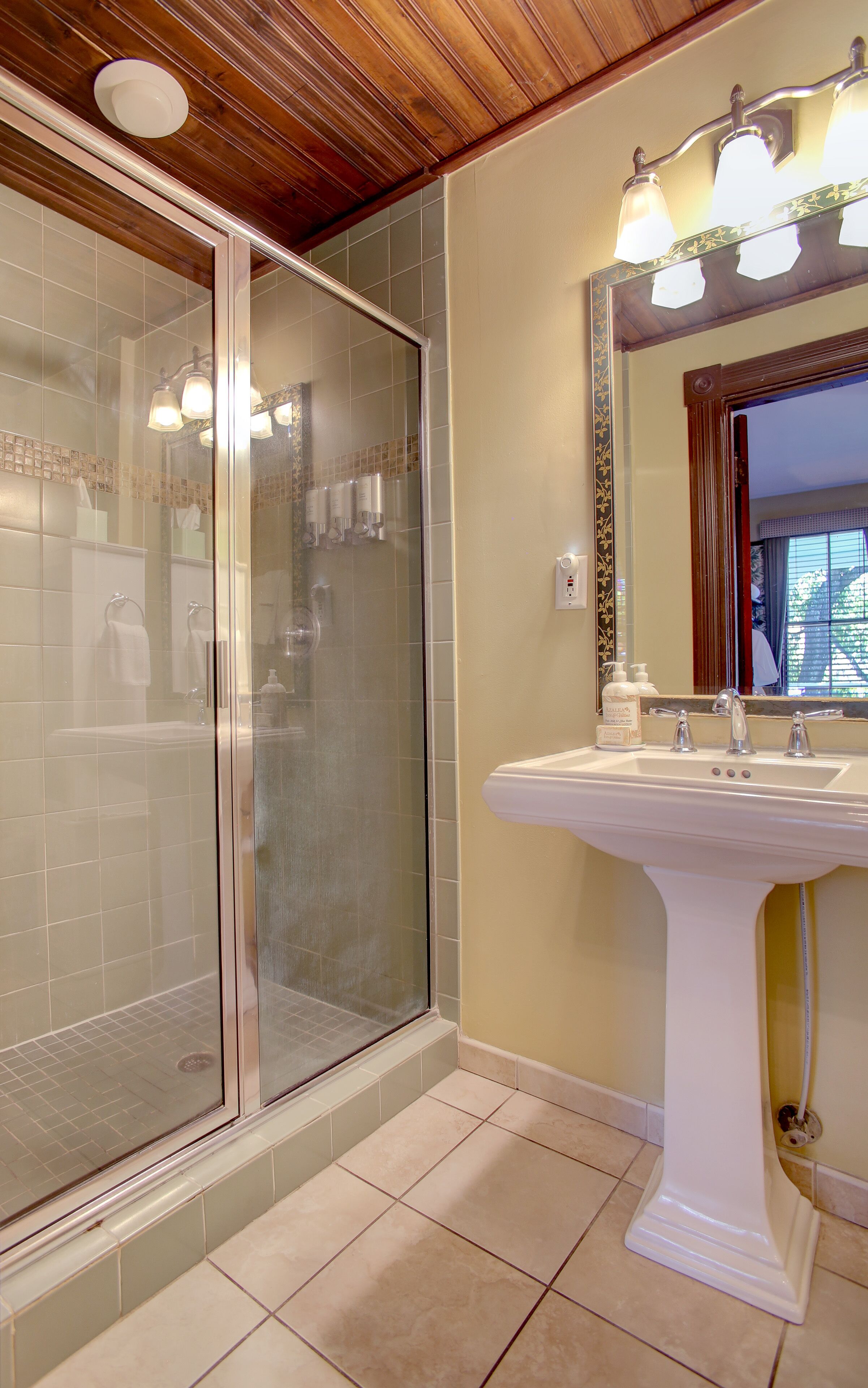 A bathroom with a glass-enclosed shower, a white sink with three faucets, and a mirror with a decorative frame.