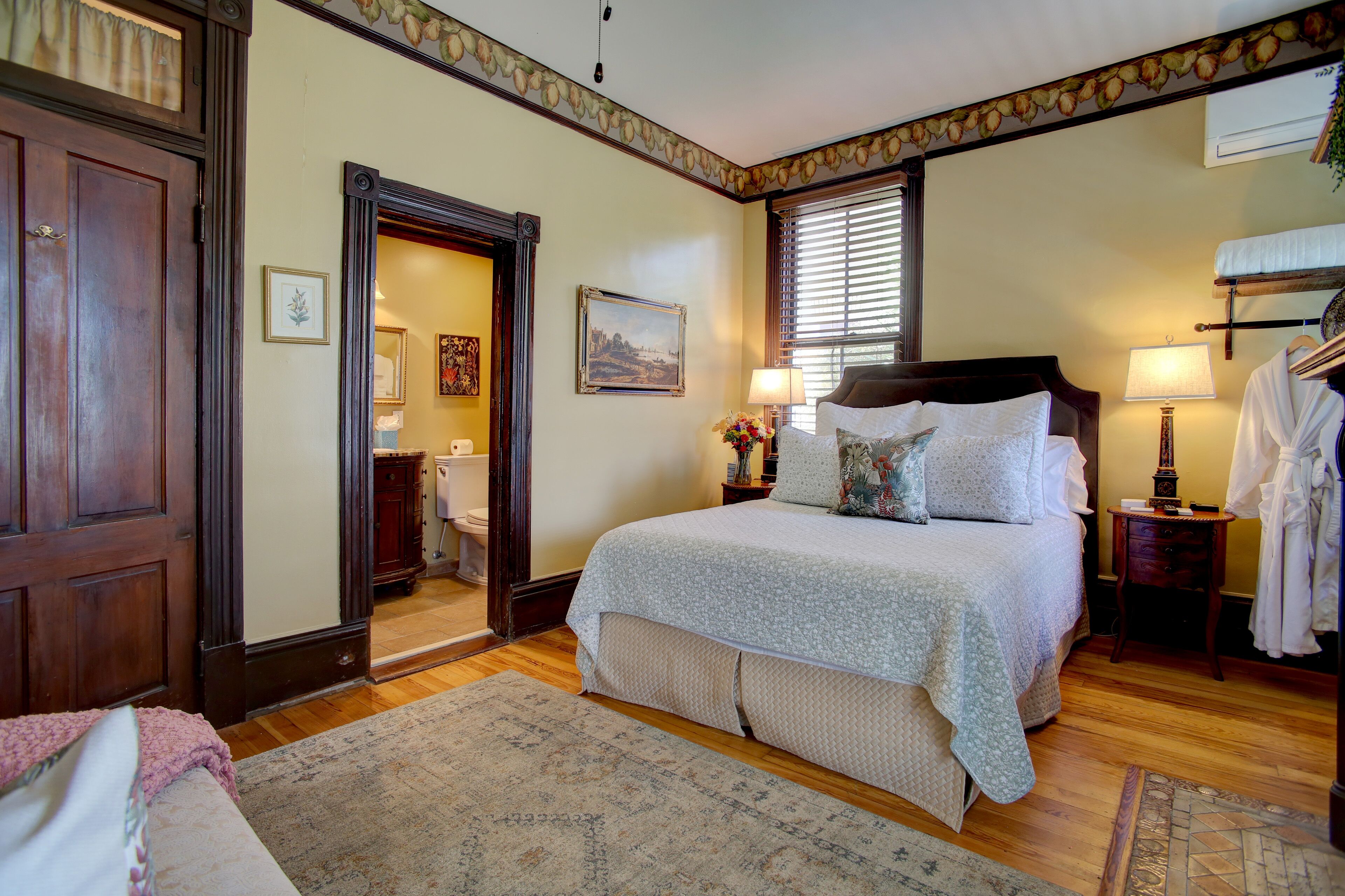A bedroom with a large bed, wooden flooring, a door leading to a bathroom, and a small table with a lamp.