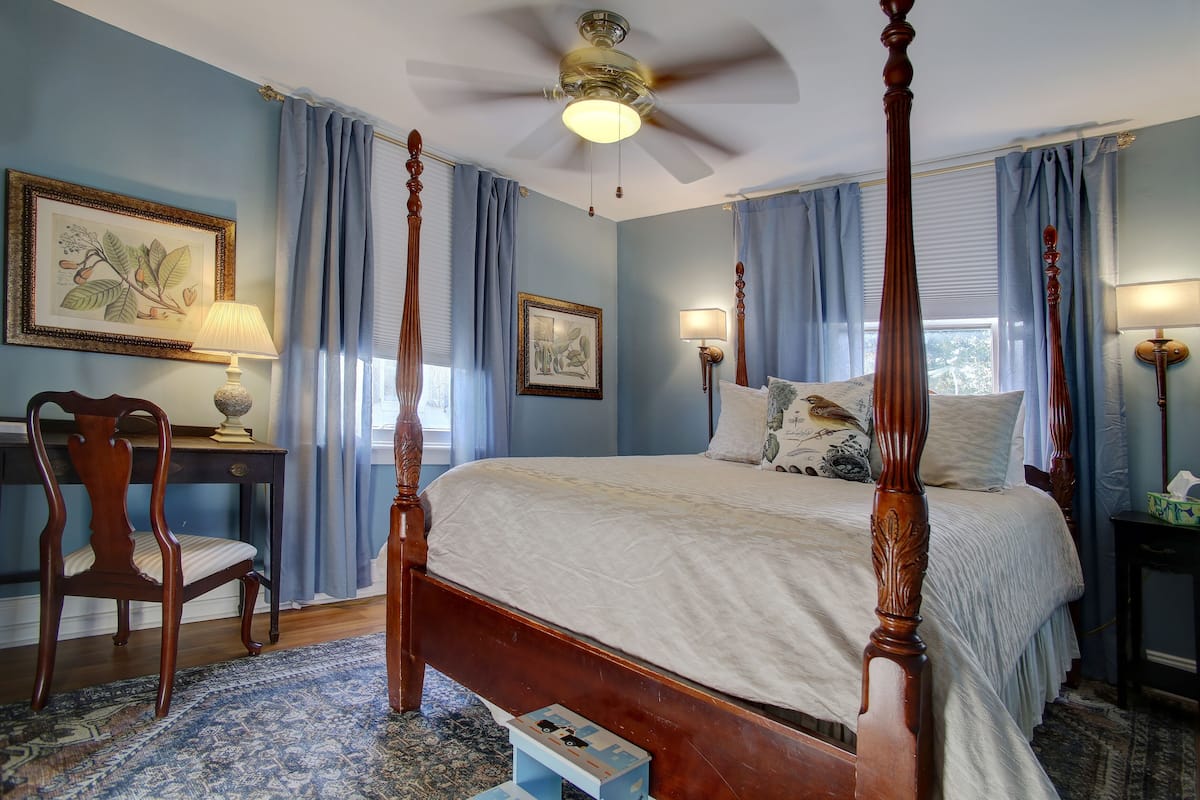 A four-poster bed with a wooden frame, a desk with a lamp, and a chair in a room with blue walls and curtains.