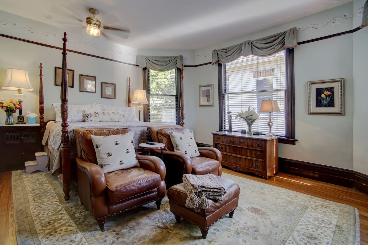 A bedroom with a four-poster bed, two leather armchairs, a wooden dresser, and a large window with curtains.