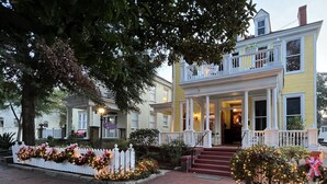 A two-story yellow house with a porch, white picket fence, and Christmas lights.