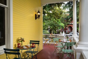 A yellow exterior wall with a white door, a small table with chairs, and a white railing with a view of trees and buildings. - Azalea Inn and Villas (Savannah)