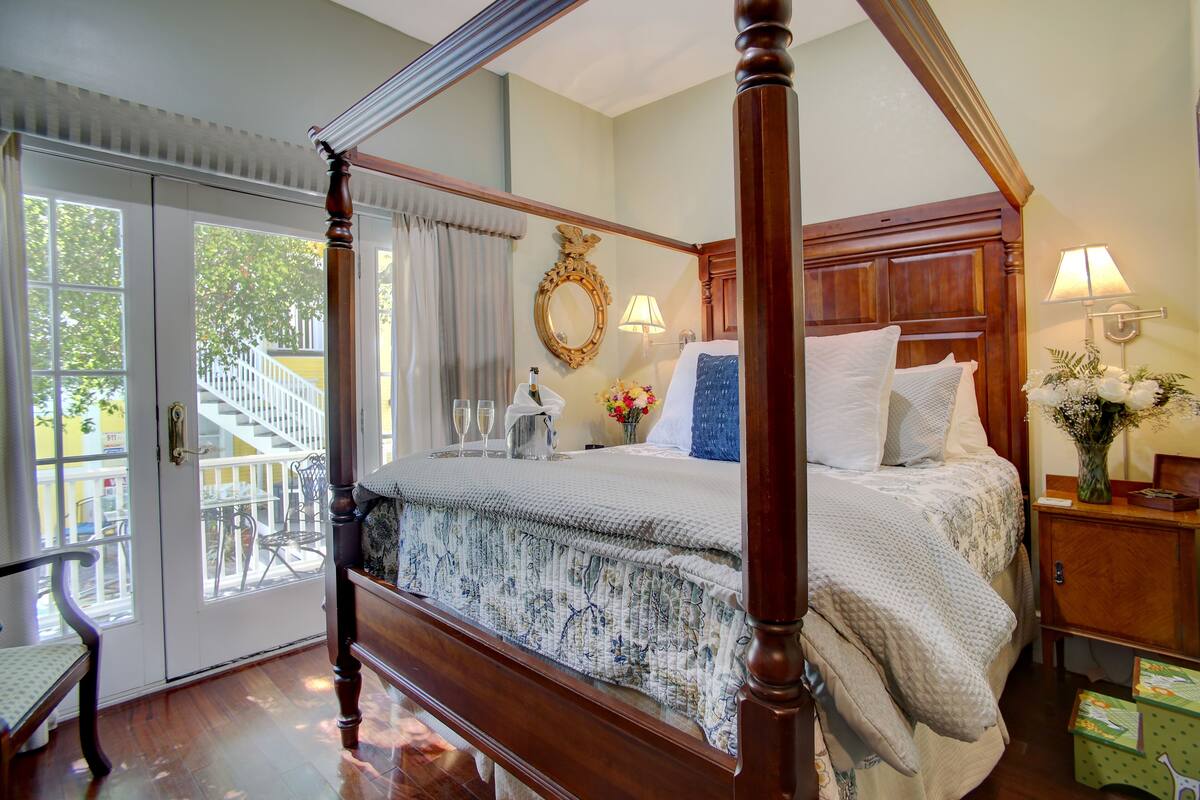 A four-poster bed with a floral bedspread, a nightstand with a lamp, and a view of a balcony with chairs.